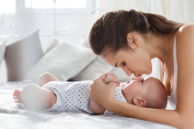 Genç anne yeni doğmuş bebeğiyle yatakta uzanıyor, birlikte oynuyor, mutluluk aile kavramı, çocuk ve anne bağı. Güzel genç anne çocuğuna bakıyor ve gülümsüyor, yatakta uzanıyor.