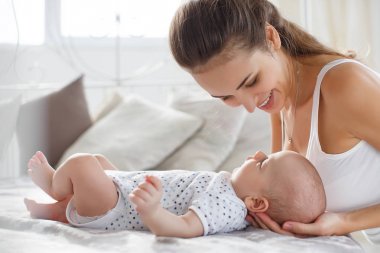 Genç anne yeni doğmuş bebeğiyle yatakta uzanıyor, birlikte oynuyor, mutluluk aile kavramı, çocuk ve anne bağı. Güzel genç anne çocuğuna bakıyor ve gülümsüyor, yatakta uzanıyor.