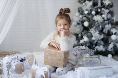 Mutlu çocuk, 5 yaşındaki kız yakınındaki beyaz oyuncaklar ve kar taneleri, dekore edilmiş güzel yeşil Noel ağacı, Noel tatili oturur güzel Noel hediyeleri beyaz bir katta büyük kutularındaki yanında mutlu çocukluk