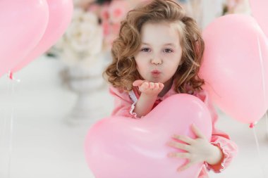 6 yaş pembe elbise ve beyaz tayt, çok güzel bir gülümsemen giymiş uzun kıvırcık saçlı küçük bir kız yalnız bir kalp şeklinde pembe balon dolu bir büyük parlak odasında oturuyor. Sevgililer günü ve parti kutlama.