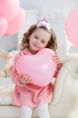 6 yaş pembe elbise ve beyaz tayt, çok güzel bir gülümsemen giymiş uzun kıvırcık saçlı küçük bir kız yalnız bir kalp şeklinde pembe balon dolu bir büyük parlak odasında oturuyor. Sevgililer günü ve parti kutlama.