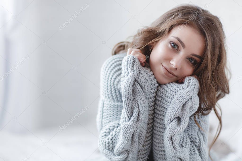 Winter Concept. Cute young girl in a gray knitted sweater. Beautiful woman is relaxing in a white bedroom. Beautiful women in winter clothes are waking up in the morning. Woman wearing a sweater in a white bedroom.