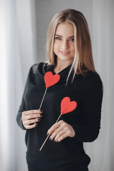 Young beautiful woman with a nice smile and big grey eyes,a blonde with long straight hair,dressed in a black party dress, spends time alone in the big bright room,holding hands of little Krasnye the heart is a symbol of Valentine\'s day