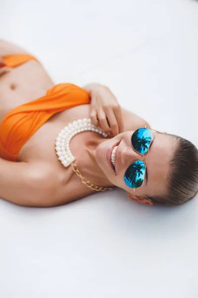 Elegant sexy woman in bikini on sun-tanned slim and shapely body posing near swimming pool. Gorgeous young woman posing in orange bikini lying by the pool in the mirror-blue glasses.