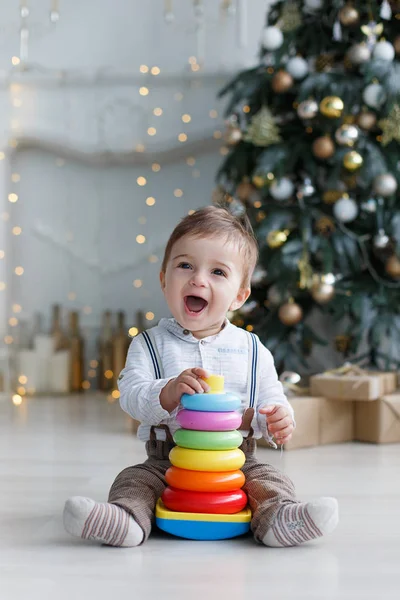 一个小男孩坐在白色光泽的地板附近的一个聪明的 绿色的圣诞树与礼品盒下 在节日装饰和灯光的背景下 一个收集一个多色塑料金字塔 — 图库照片