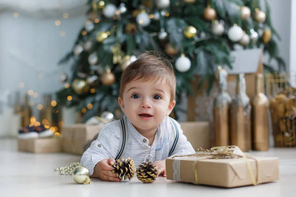 棕色的大眼睛 金色短发控股圣诞树金颜色的玩具的小男孩穿着一件棕色的裤子和白色的条纹的衬衫 坐在旁边一棵白色的圣诞树和礼品包装盒的地板上 — 图库照片