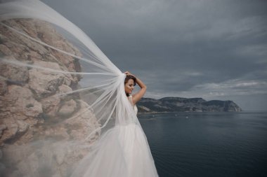 Güzel gelin peçe bir göz alıcı beyaz düğün elbise görünümünde deniz cliff üzerinde duruyor. Romantik arka plan denizin üzerinde poz beyaz elbiseli güzel gelin. Sahilde poz lüks elbiseli mutlu gelin. Düğün günün kutlu olsun.