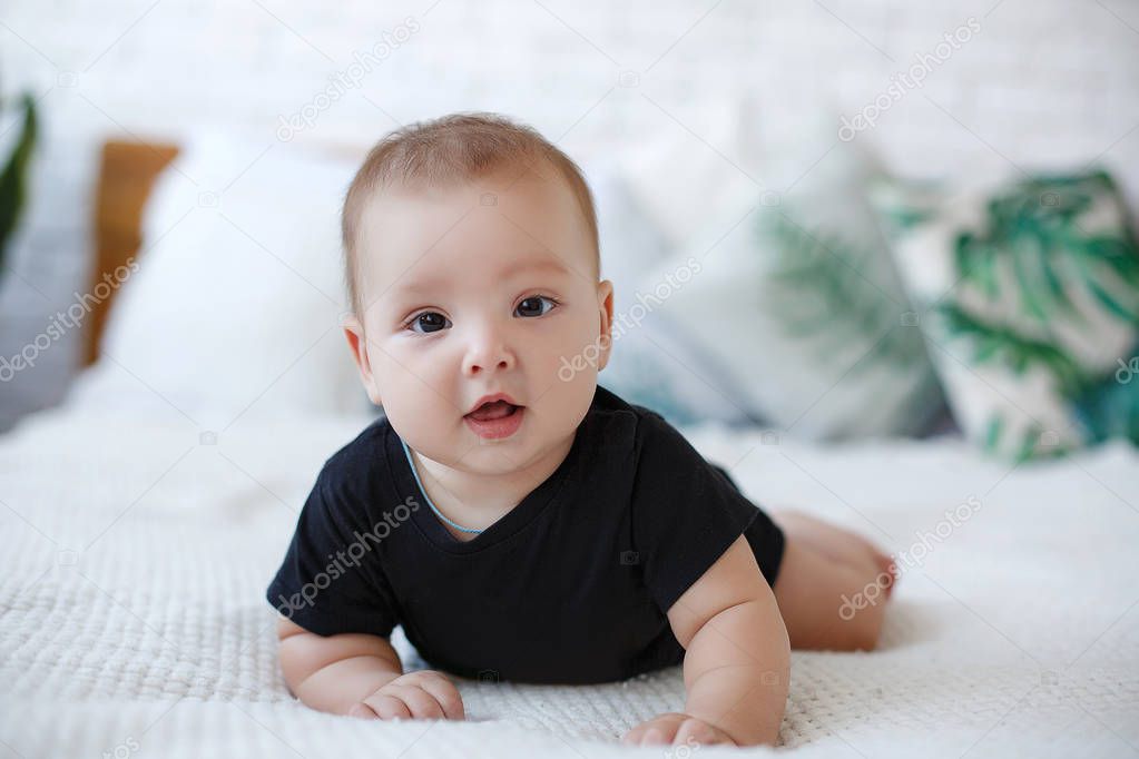 Adorable baby boy in white sunny bedroom plays alone lying  on the bed.Charming beautiful lovely small female baby with plump cheeks creeps on bed, looks innocently on something, being naughty. Cute baby infant plays alone. Newborn kid crawls.