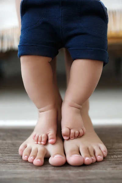 The legs of the baby stand on the feet of the mother, the mother supports the little son.Adorable baby\'s and mother\'s feet on floor at living room. Legs of mother and baby. First steps. Mom teaches her child to stand and de balance