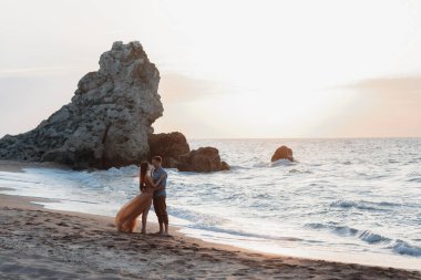 Deniz kenarında şafak vakti aşık bir çift. Balayı gezisi. Erkek ve kadın seyahat ediyor. Deniz manzaralı mutlu bir çift. Erkek ve kadın el ele tutuşuyor. Tatilde aşık bir çift. Beni takip edin. Güneşli bir günde, mavi okyanusun kayalık sahillerinde bir çift