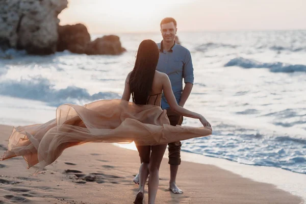 Couple in love at dawn by the sea. Honeymoon trip. Man and woman traveling. Happy couple by the sea view from the back. Man and woman holding hands. Couple in love on vacation. Follow me. Couple on a rocky beach of blue ocean on a sunny day