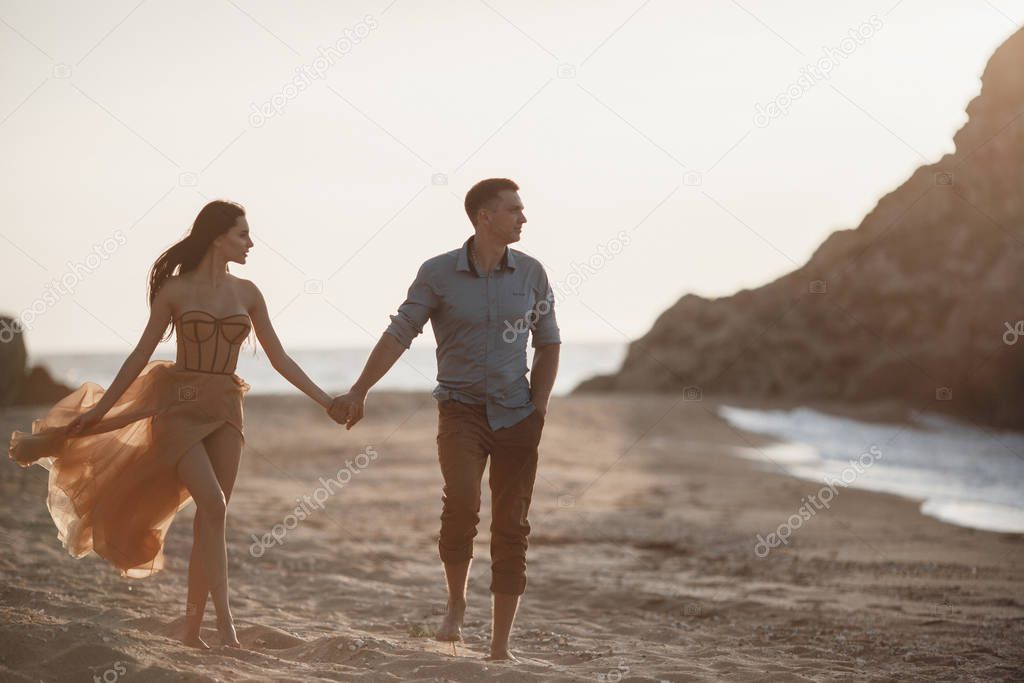 Couple in love at dawn by the sea. Honeymoon trip. Man and woman traveling. Happy couple by the sea view from the back. Man and woman holding hands. Couple in love on vacation. Follow me. Couple on a rocky beach of blue ocean on a sunny day