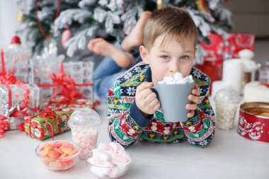 Noel arifesinde elinde bir bardak milkshake olan çocuk evde Noel ağacının yanında yatıyor. 7-8 yaşlarında tatlı bir çocuk, elinde bir bardakla şık bir Noel ağacının yanında yatıyor. Kış süslemeli, aydınlık bir yatak odasında. Noel ve yeni yıl tatili, Aralık..