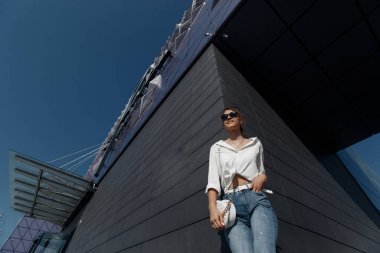 Kot pantolonlu, açık havada poz veren uzun saçlı esmer bir kadın. Moda fotoğrafı konsepti. İş merkezine yakın bir kadın. Esmer bir kadının portresi. Ofisinin yanında güzel bir kadın iş merkezi çalışanı.
