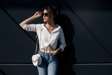 Kot pantolonlu, açık havada poz veren uzun saçlı esmer bir kadın. Moda fotoğrafı konsepti. İş merkezine yakın bir kadın. Esmer bir kadının portresi. Ofisinin yanında güzel bir kadın iş merkezi çalışanı.