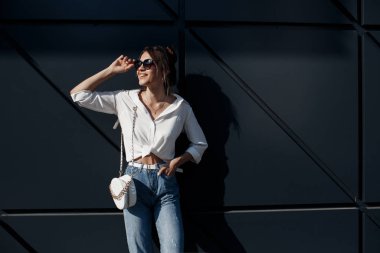 Kot pantolonlu, açık havada poz veren uzun saçlı esmer bir kadın. Moda fotoğrafı konsepti. İş merkezine yakın bir kadın. Esmer bir kadının portresi. Ofisinin yanında güzel bir kadın iş merkezi çalışanı.