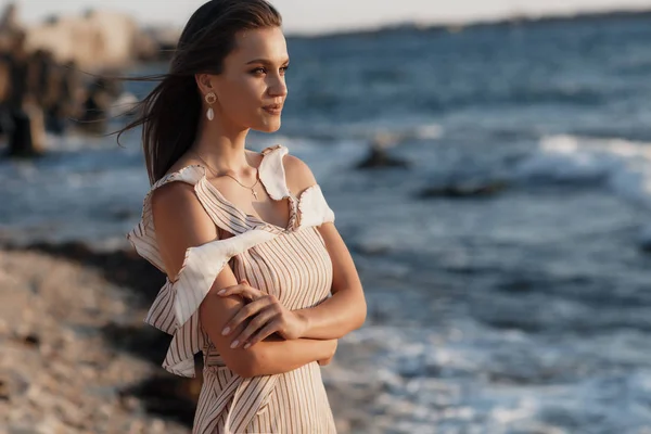 Beautiful young woman posing against the sea and skyline.Summer vacation concept. Happy woman relaxing near the sea on beach.Hipster slim girl in beige dress standing and tanning on beach near sea with waves,sunny warm weather. Peaceful calm moment.