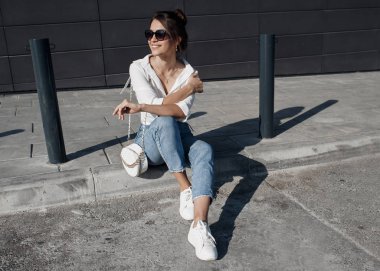 Kot pantolonlu, açık havada poz veren uzun saçlı esmer bir kadın. Moda fotoğrafı konsepti. İş merkezine yakın bir kadın. Esmer bir kadının portresi. Ofisinin yanında güzel bir kadın iş merkezi çalışanı.