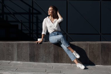 Kot pantolonlu, açık havada poz veren uzun saçlı esmer bir kadın. Moda fotoğrafı konsepti. İş merkezine yakın bir kadın. Esmer bir kadının portresi. Ofisinin yanında güzel bir kadın iş merkezi çalışanı.
