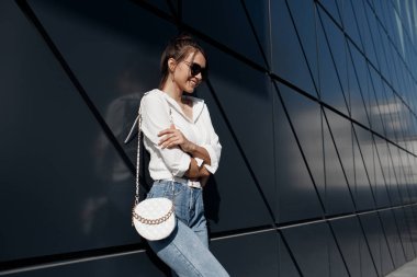 Kot pantolonlu, açık havada poz veren uzun saçlı esmer bir kadın. Moda fotoğrafı konsepti. İş merkezine yakın bir kadın. Esmer bir kadının portresi. Ofisinin yanında güzel bir kadın iş merkezi çalışanı.