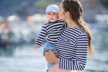 Mom and son are sitting on the old pier and enjoying the sea.Happy mother and baby in striped navy t-shirts spend time together in the summer in the summer on a wooden pier near the sea and boats. Warm sunny summer day.