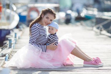 Mom and son are sitting on the old pier and enjoying the sea.Happy mother and baby in striped navy t-shirts spend time together in the summer in the summer on a wooden pier near the sea and boats. Warm sunny summer day.