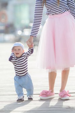 Mom and son are sitting on the old pier and enjoying the sea.Happy mother and baby in striped navy t-shirts spend time together in the summer in the summer on a wooden pier near the sea and boats. Warm sunny summer day.