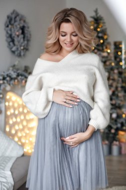 Genç ve güzel hamile bir kadın gülümsüyor ve mutlu görünüyor, saç şekillendirici makyaj, arka planda Noel ağacı ve şömine rafında beyaz gümüş rengi. Noel ağacının yanındaki güzel hamile kadının portresi..