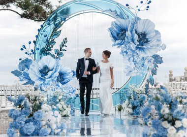 Açık hava şölen salonunda büyük mavi çiçeklerle süslenmiş bir düğün töreni. Gelin ve damat birbirlerinin ellerini tutuyorlar. Düğün günü bir yaz restoranında güzel dekorasyonu ve tasarımları olan bir ziyafet. Aşık mutlu genç bir çift.
