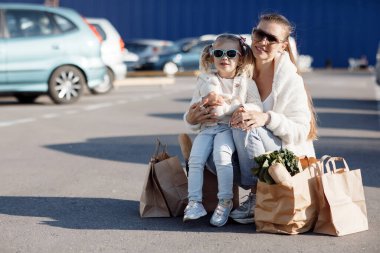 Anne ve küçük kızı at kuyruklu, beyaz tişörtlü ve kot pantolonlu süpermarketin yanındaki otoparkta ellerinde paketlerle poz veriyorlar. Anne ve kızı dışarıda alışveriş yapıyor..