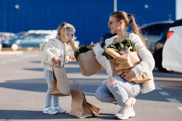 妈妈和小女儿 戴着马尾辫 穿着白色T恤衫和蓝色牛仔裤 站在超市附近停车场的后面 手里拿着包裹 母亲和女儿在户外购物 — 图库照片