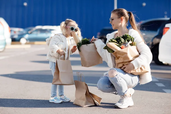 白いTシャツを着たポニーテールのママと小さな娘 スーパーの近くの駐車場の背景にポーズをとった青いジーンズ 母親と娘が屋外で買い物 — ストック写真
