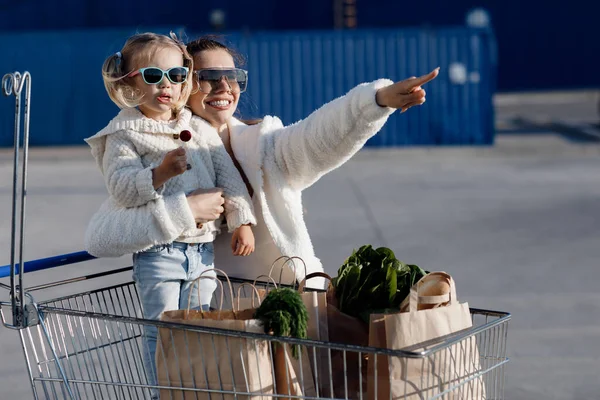 妈妈和小女儿 戴着马尾辫 穿着白色T恤衫和蓝色牛仔裤 站在超市附近停车场的后面 手里拿着包裹 母亲和女儿在户外购物 — 图库照片