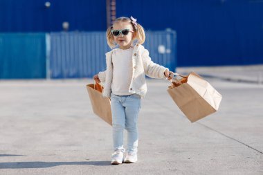 Alımlı neşeli bir bebek. Alışveriş torbaları tutan küçük bir kız. Baharda alışveriş torbalarıyla yalnız başına. Güneş gözlüklü güzel bir kız, alışveriş torbalarıyla sokakta yalnız poz veriyor.