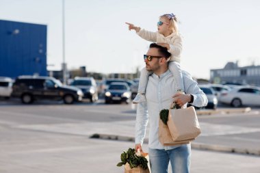 Beyaz bir baba, küçük kızıyla marketten alışveriş yapıyor. Babam taze sebze alıyor. Küçük kızı olan mutlu bir baba otoparkta elinde taze sebzeler ve alışveriş torbalarıyla yürüyor..
