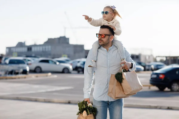 Caucasian father shopping in grocery store with baby daughter. Dad buying fresh vegetables. A happy father with a small daughter on his shoulders is walking in the parking lot holding paper bags in his hands with fresh vegetables and shopping.