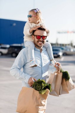 Beyaz bir baba, küçük kızıyla marketten alışveriş yapıyor. Babam taze sebze alıyor. Küçük kızı olan mutlu bir baba otoparkta elinde taze sebzeler ve alışveriş torbalarıyla yürüyor..