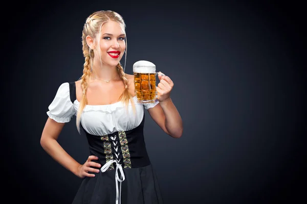 Joven camarera sexy Oktoberfest, con un vestido tradicional bávaro, sirviendo tazas de cerveza grandes sobre fondo negro . — Foto de Stock