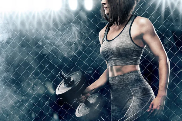 Brutal athletic woman pumping up muscles with dumbbells on black background with smoke — Stock Photo, Image