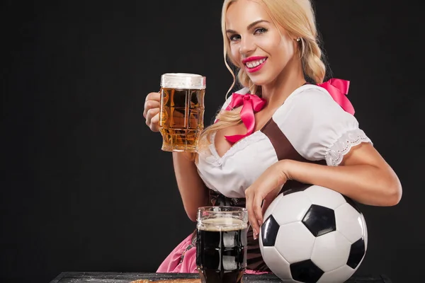 Junges sexy Oktoberfestmädchen - Kellnerin in traditioneller bayerischer Tracht, serviert große Bierkrüge und nimmt Fußballball auf schwarzem Hintergrund. — Stockfoto