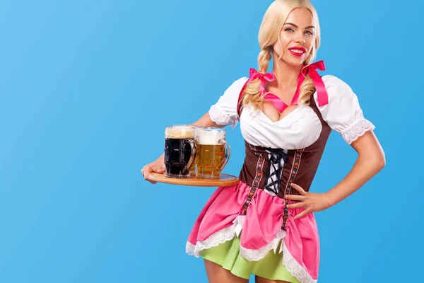 Jonge sexy Oktoberfest meisje - serveerster, dragen een traditionele Beierse kleding, serveren van grote bierpullen op blauwe achtergrond. — Stockfoto