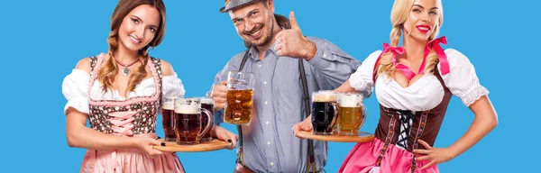 Portret van gelukkig Oktoberfest man en twee vrouwen, het dragen van een traditionele Beierse kleding, ten dienste van groot bier mokken op blauwe achtergrond. — Stockfoto