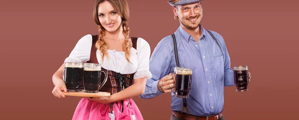 Portret van gelukkig Oktoberfest man en vrouw, het dragen van een traditionele Beierse kleding, serveren van grote bierpullen op bruine achtergrond. — Stockfoto