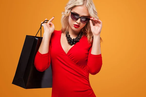 Blond kvinna i solglasögon på shopping håller mörk väska på orange bakgrund på svart fredag semester. Kopiera utrymme för salu annonser. — Stockfoto