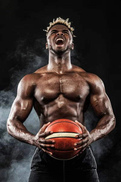 Portrait d'un sportif afro-américain, joueur de basket avec une balle sur fond noir. S'adapter jeune homme en tenue de sport ballon . — Photo