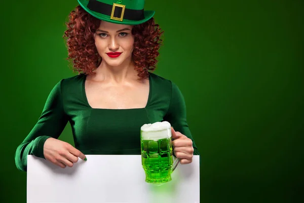 St Patrick Day redhead girl with beer mug. Young sexy oktoberfest woman, wearing a dress and green hat with white banner for copy space. — Stock Photo, Image