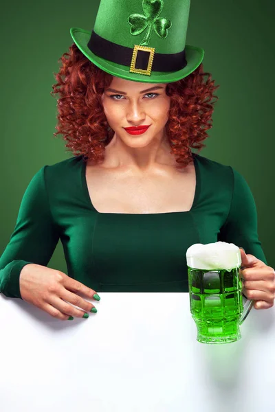 St Patrick Day rödhårig tjej med ölglas. Ung sexig oktoberfest kvinna, som bär en klänning och grön hatt med vita banner för kopia utrymme. — Stockfoto