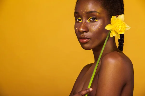 Cheerful young african woman with yellow makeup on her eyes. Female model against yellow background with yellow flower.