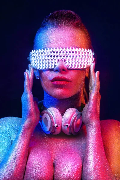 O femeie frumoasă, cu sclipici și străluciri pe față și pe corp. Portret de TDJ sexy cu căști și ochelari de soare neon — Fotografie, imagine de stoc