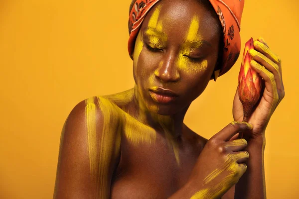 Vrolijke jonge Afrikaanse vrouw met gele make-up op haar ogen. Vrouwelijke model tegen een gele achtergrond met oranje bloem. — Stockfoto